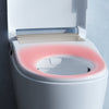 A close-up of a white toilet with the lid open, showing a pink illuminated seat surround, set against a gray wall with a soft shadow.