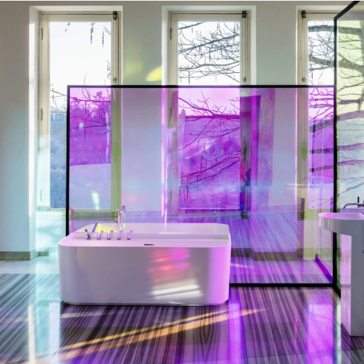 A modern bathroom interior with a freestanding white bathtub and matching white pedestal sink, separated by a translucent purple-tinted glass screen. Large windows behind show bare winter trees, and the floor reflects the purple light cast across the room.